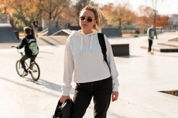 Outdoor portrait of young beautiful woman with a ponytail and sunglasses, with a backpack on his shoulders, dressed in a white sweater, near the sportsground. White hoodie