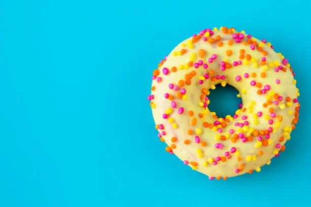 Yellow donut on a blue background with copy space. Multicolor topping on yellow glaze. Unhealthy high-calorie food