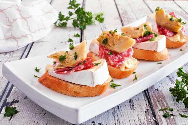 aperitivos de crostini con queso brie, salami y alcachofas, cerca de un plato contra la madera blanca - cheese still life tray french cuisine fotografías e imágenes de stock