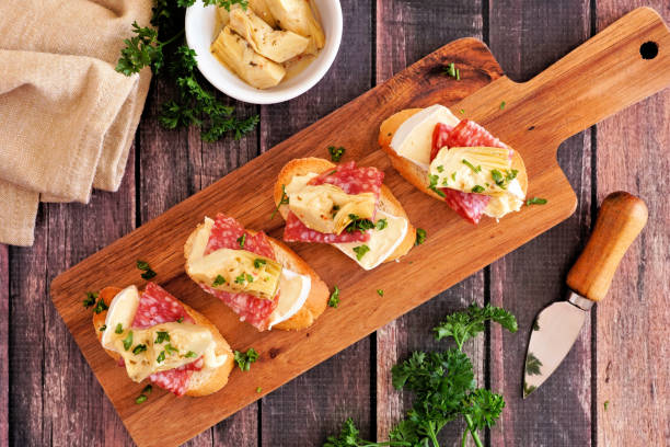 aperitivos crostini con queso brie, salami y alcachofas, vista superior en una tabla de servicio contra madera rústica - cheese still life tray french cuisine fotografías e imágenes de stock