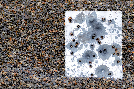 A piece of metal filled with bullet holes. The metal is resting in a pile of dark wet gravel and appears to be the back of a sign. Lots of bullet holes. Room for text.