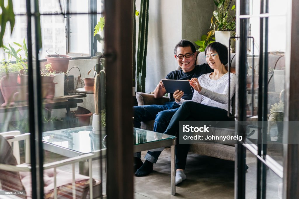 Cheerful Chinese woman showing man digital tablet in conservatory Couple sitting on sofa at home using device, woman touching screen and smiling, view through open door Asian and Indian Ethnicities Stock Photo