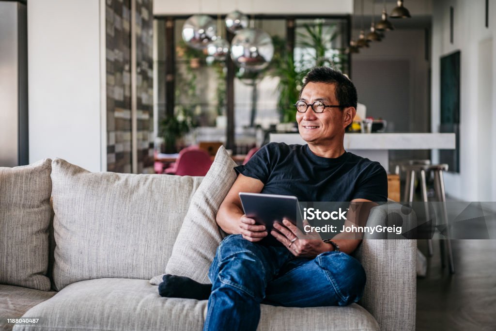 Cheerful Chinese man with tablet looking away and smiling Man in his 50s sitting on sofa at home, leisure time, relaxation, enjoyment Men Stock Photo