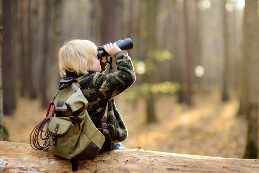Little boy scout with binoculars during hiking in autumn forest. Child is sitting on large fallen tree and looking through a binoculars. Concepts of adventure, scouting and hiking tourism for kids.