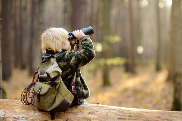 mały harcerz z lornetką podczas wędrówki po jesiennym lesie. dziecko siedzi na dużym powalonym drzewie i patrzy przez lornetkę. - tree skill nature horizontal zdjęcia i obrazy z banku zdjęć