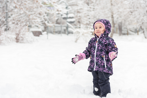 Happy baby girl rejoices in falling snow, Moscow, Russia. Cute little child having fun plays in snowy park. Adorable smiling kid walks during snowfall. Joyful child outside in winter for background.