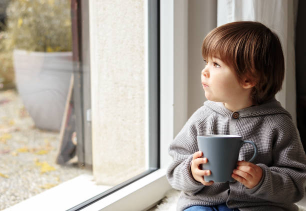 menino pequeno bonito da criança que senta-se no indicador com copo do chá morno que olha para fora pensativamente no tempo frio do outono. casa acolhedora. queda conceito melancólico. humor sazonal - child autumn nature human face - fotografias e filmes do acervo