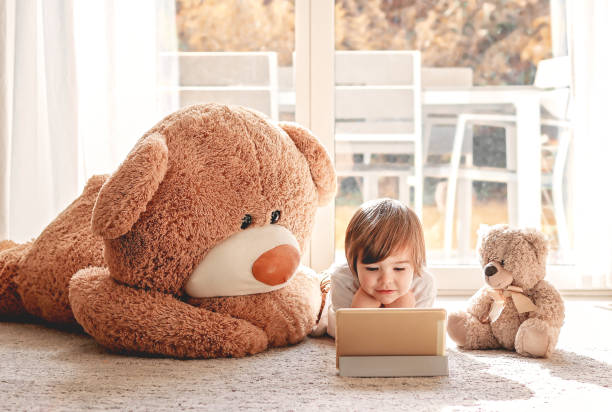 Cute little child watching cartoons on digital tablet device lying on floor with two soft teddy bear toys at home. Modern childhood. stock photo