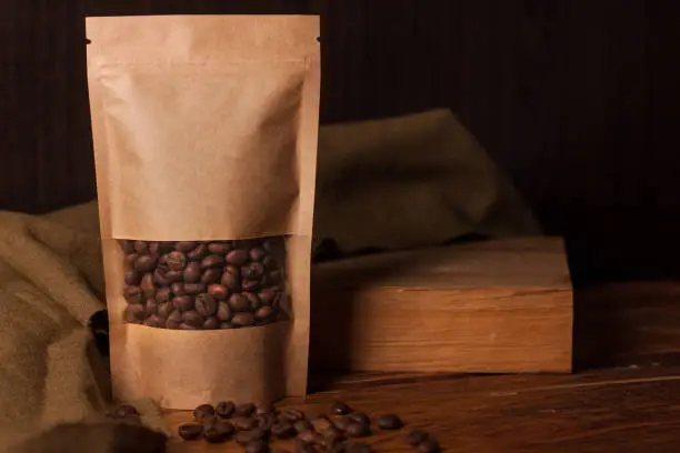 Photo of Blank brown kraft paper pouch bag with coffee beans in transparent window on wooden background. Trendy packaging for tea