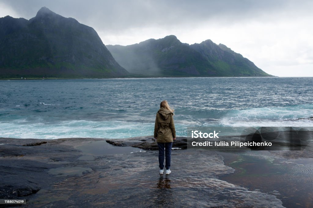 Blond hair girl with a backpack stands on big stones near the water and looks at the ocean. Waves, splashing. Enjoy the moment, relaxation. Wanderlust. Travel, adventure, lifestyle. Explore Norway Sea Stock Photo
