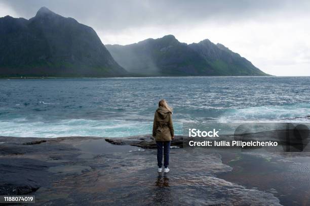 Blond Hair Girl With A Backpack Stands On Big Stones Near The Water And Looks At The Ocean Waves Splashing Enjoy The Moment Relaxation Wanderlust Travel Adventure Lifestyle Explore Norway Stock Photo - Download Image Now