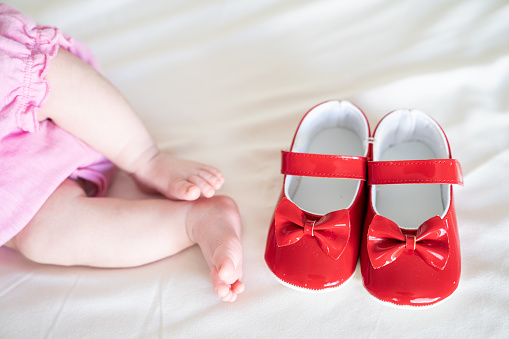 Close up photo of baby foot and brand new red first shoes on bed. Only foots next to shoes are in view. Shot under daylight with a full frame mirrorless camera.