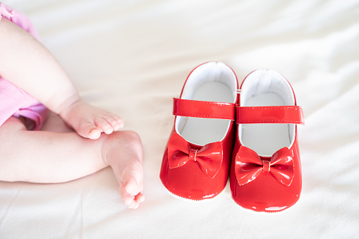 Close up photo of baby foot and brand new red first shoes on bed. Only foots next to shoes are in view. Shot under daylight with a full frame mirrorless camera.
