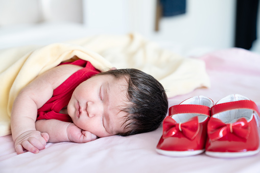 Portrait of sleeping newborn baby girl dreaming of walking. Her first cute red shoes are seen next to her. Her eyes are closed. Shot under daylight with a full frame mirrorless camera.