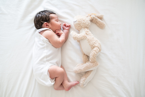 High angle full length portrait of newborn baby girl sleeping with stuffed toy rabbit on bed. Shot indoor with a full frame mirrorless camera.