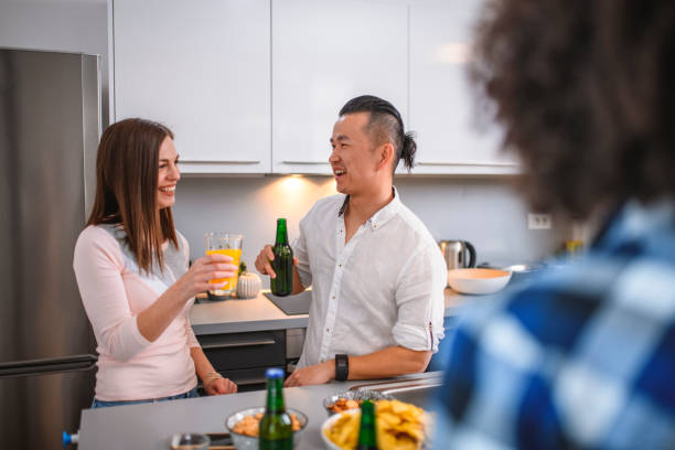 Asian and Caucasian Friends Talking and Flirting at Party Over the shoulder view of young Asian male and Caucasian female enjoying drinks and conversation in kitchen at social gathering. half shaved hairstyle stock pictures, royalty-free photos & images