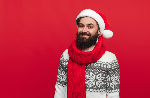 Cheerful bearded hipster in Santa hat and knitted clothes smiling and looking at camera while standing against red background during Christmas celebration