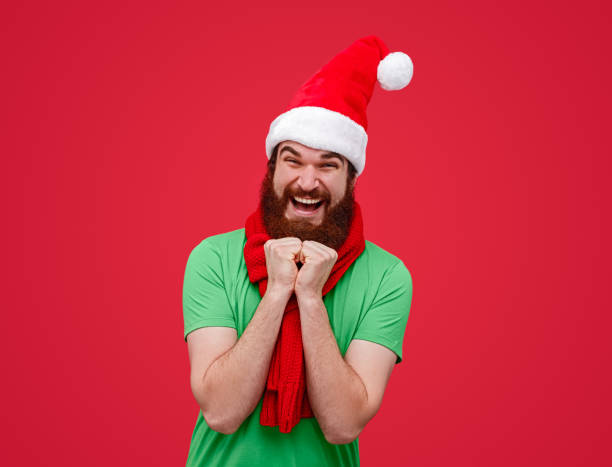 Excited Santa assistant looking at camera Amazed bearded elf in cap and scarf clasping hands and looking at camera against red background on Christmas Day christmas excitement stock pictures, royalty-free photos & images