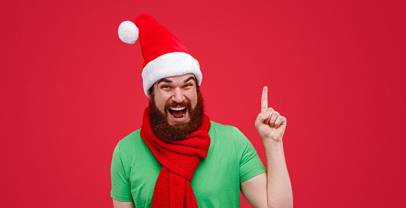 Happy bearded man in elf costume looking at camera and pointing up during Christmas celebration against red background