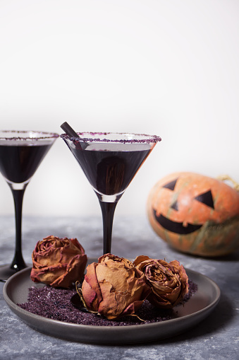 Two glasses with black cocktail, dried roses and Jack-o'-lantern for Halloween party on the dark background.
