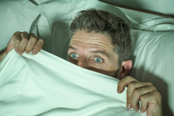 ritratto drammatico di uomo stressato e spaventato da solo a letto sveglio di notte nella paura dopo aver avuto un incubo sensazione paranoica tenendo la coperta in divertente espressione faccia di panico "n - attractive male men humor looking at camera foto e immagini stock