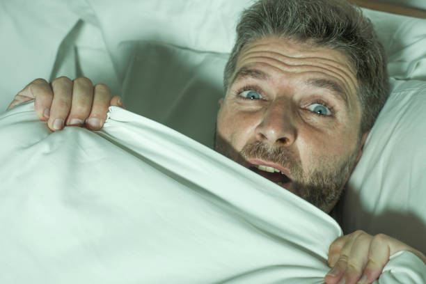 ritratto drammatico di uomo stressato e spaventato da solo a letto sveglio di notte nella paura dopo aver avuto un incubo sensazione paranoica tenendo la coperta in divertente espressione faccia di panico "n - attractive male men humor looking at camera foto e immagini stock