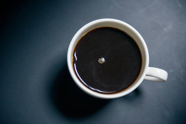 Detailed close-up from above of an old-fashioned white porcelain mug filled with freshly poured black coffee, with a bit of foam on top. the background is white with a soft shadow gradient to the right. the handle is on the left