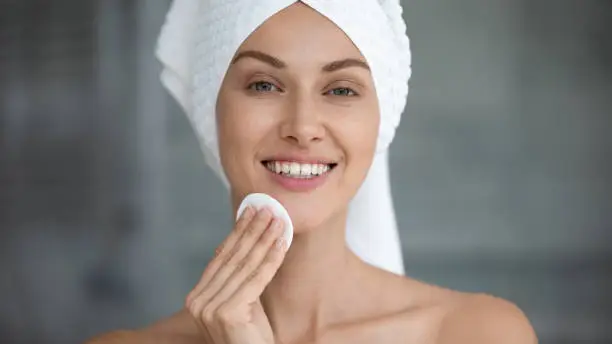 Smiling beautiful young woman with towel on head hold cotton pad disk cleansing face skin with cleanser, happy lady remove makeup look at camera enjoy healthy clean skincare beauty treatment concept