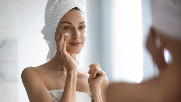 Attractive young adult woman applying facial cream looking in mirror Attractive young adult woman apply facial cream look in mirror, beautiful healthy lady wrapped in towels put moisturizing lifting nourishing day creme on soft hydrated moisturized skin in bathroom moisturizing cream stock pictures, royalty-free photos & images