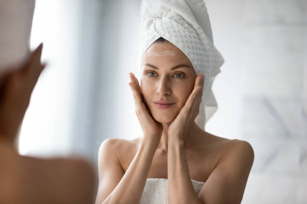 красивая молодая женщина смотреть в зеркало массаж лица применения крема - human skin aging process beautiful hygiene стоковые фото и изображения