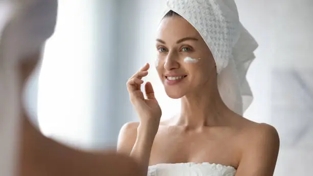 Photo of Smiling pretty lady put moisturizing facial cream look in mirror