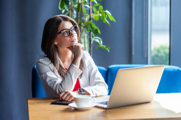 verticale de la jeune femme élégante réfléchie de brunette dans des lunettes s'asseyant, regardant loin et planifiant la nouvelle idée ou pensant à quelque chose nouveau. - business slave photos et images de collection