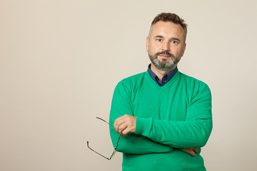 Studio portrait of a 50 year old bearded man in a green sweater holding glasses in his hands on a beige background