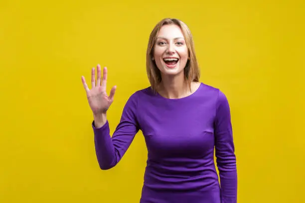 Photo of Hi! Portrait of sweet happy woman with raised hand saying hello to camera. indoor studio shot isolated on yellow background