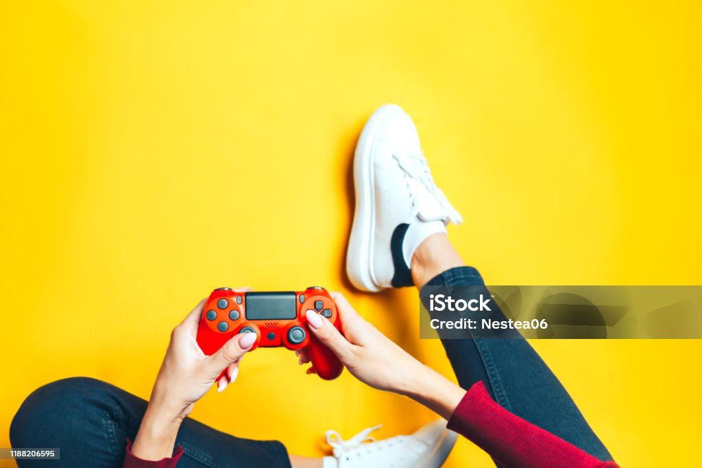 Young woman playing with two gamepads on yellow. Young thin woman playing with red gamepad, sitting on yellow background. flat lay. Video Game Stock Photo