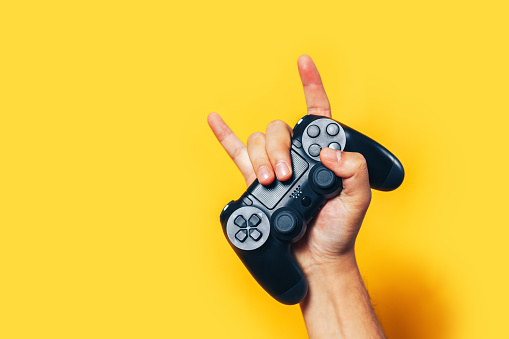 Man hand holding black gamepad show cool symbol on yellow background, minimalism concept.