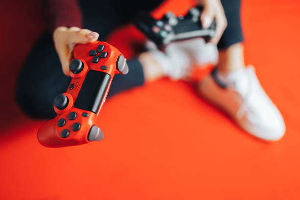 Young woman playing with two gamepads on red. Young thin woman playing with black gamepad, holding red joystick and giving to you for playing with her, sitting on red background. facilities protection services stock pictures, royalty-free photos & images