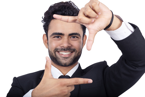 Portrait of cheerful young businessman gesturing finger frame and looking through it. Handsome Indian young male business person is on white background. Close-up of handsome man.