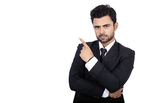 Portrait of confident Indian businessman pointing at copy space. Handsome male professional is standing on white background. He is wearing elegant suit.