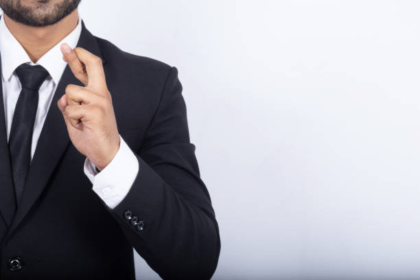 Midsection of businessman holding his fingers crossed on white background Cropped image of a businessman holding his fingers crossed. Midsection of male professional is wearing black suit. He is standing on white background asian suit man cross hand stock pictures, royalty-free photos & images