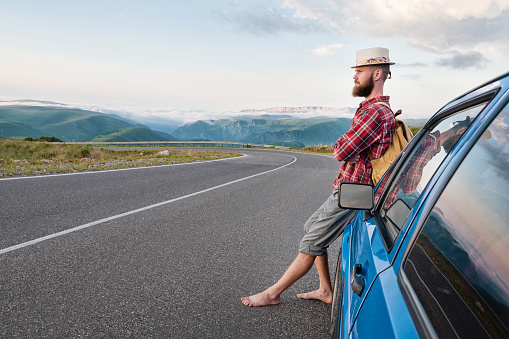 Portrait of a bearded happy traveler hipster with a backpack in a plaid shirt and a hat next to an unknown car stands on the road at sunset in the mountains. Happy and confident travel concept.
