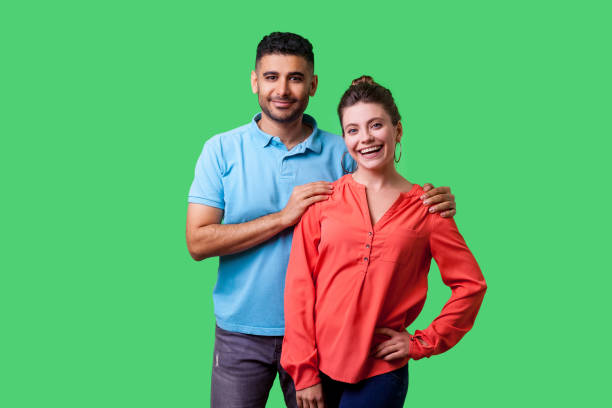 Portrait of happy attractive young couple in casual wear standing together. isolated on green background Portrait of happy attractive young couple in casual wear standing together, looking at camera with toothy smile, caring man holding female shoulders. isolated on green background, indoor studio shot hand on shoulder photos stock pictures, royalty-free photos & images