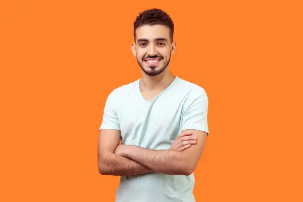Portrait of positive handsome brunette man with beard in white t-shirt standing with crossed hands and looking at camera with joyful toothy smile. indoor studio shot isolated on orange background