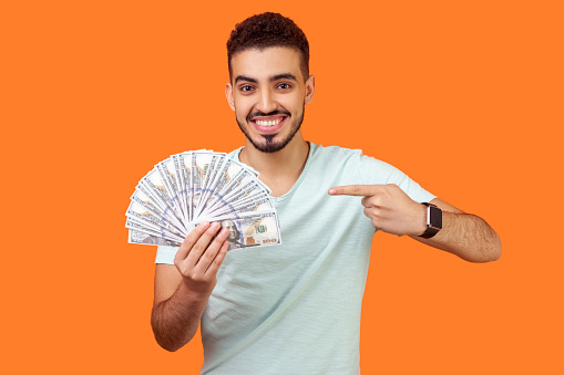 Portrait of lucky rich brunette man with beard in white t-shirt pointing at dollar banknotes and looking at camera with toothy smile, enjoying wealth. indoor studio shot isolated on orange background