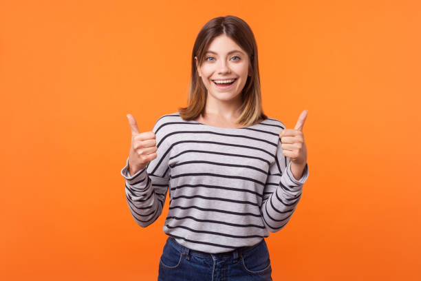 wie, daumen hoch! porträt der fröhlichen freundlichen jungen frau mit braunen haaren in langärmeligegegem hemd. isoliert auf orangefarbenem hintergrund - thumbs up human thumb human finger brown hair stock-fotos und bilder