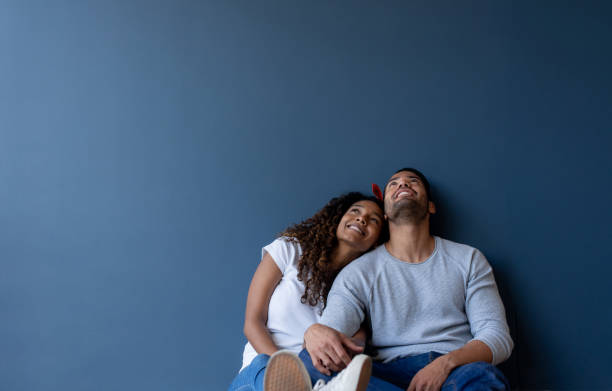 Thoughtful couple at home leaning against a wall and smiling Thoughtful couple at home leaning against a wall and smiling - real estate concepts two people thinking stock pictures, royalty-free photos & images