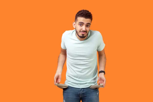 Photo of Portrait of frustrated worried brunette man turning out empty pockets. indoor studio shot isolated on orange background