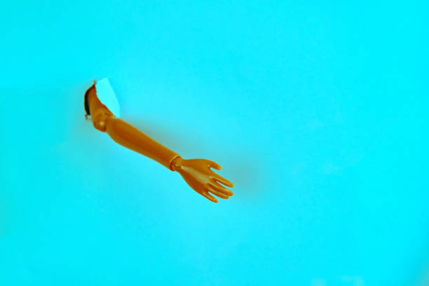hand of a plastic doll break through torn blue paper creative minimalistic still life, orange and  teal toned