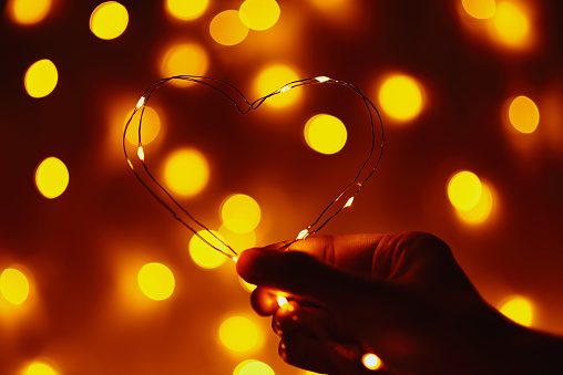 Woman hand holding wire in shape of heart against abstract background with golden blurred lights. Valentine day concept