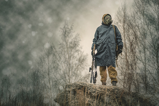 Soldier with rifle in raincoat and gas mask is walking outdoor.